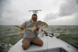 Fly angler kneeling on a Hell's Bay skiff bow holding a jack crevalle caught on fly near Pass Cavallo, Port O'Connor, Texas
